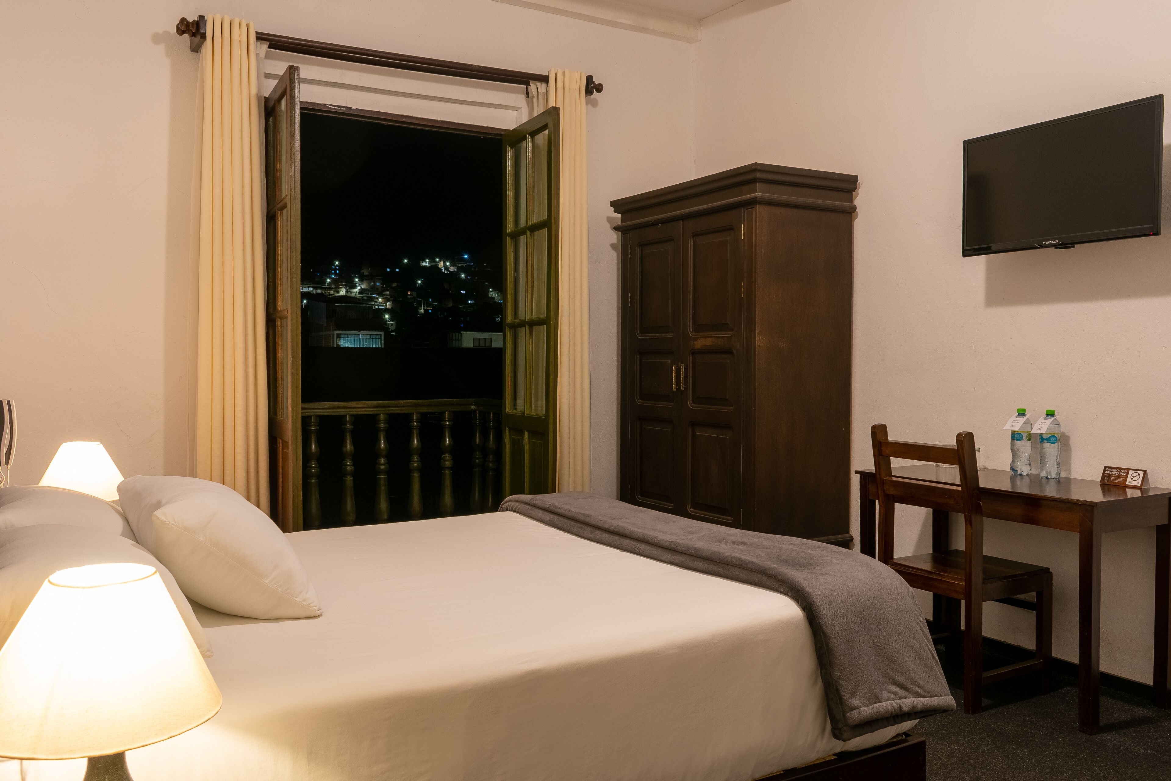 A hotel room with a bed, a desk with two bottles of water, a chair, a TV on the wall, and a window with curtains.