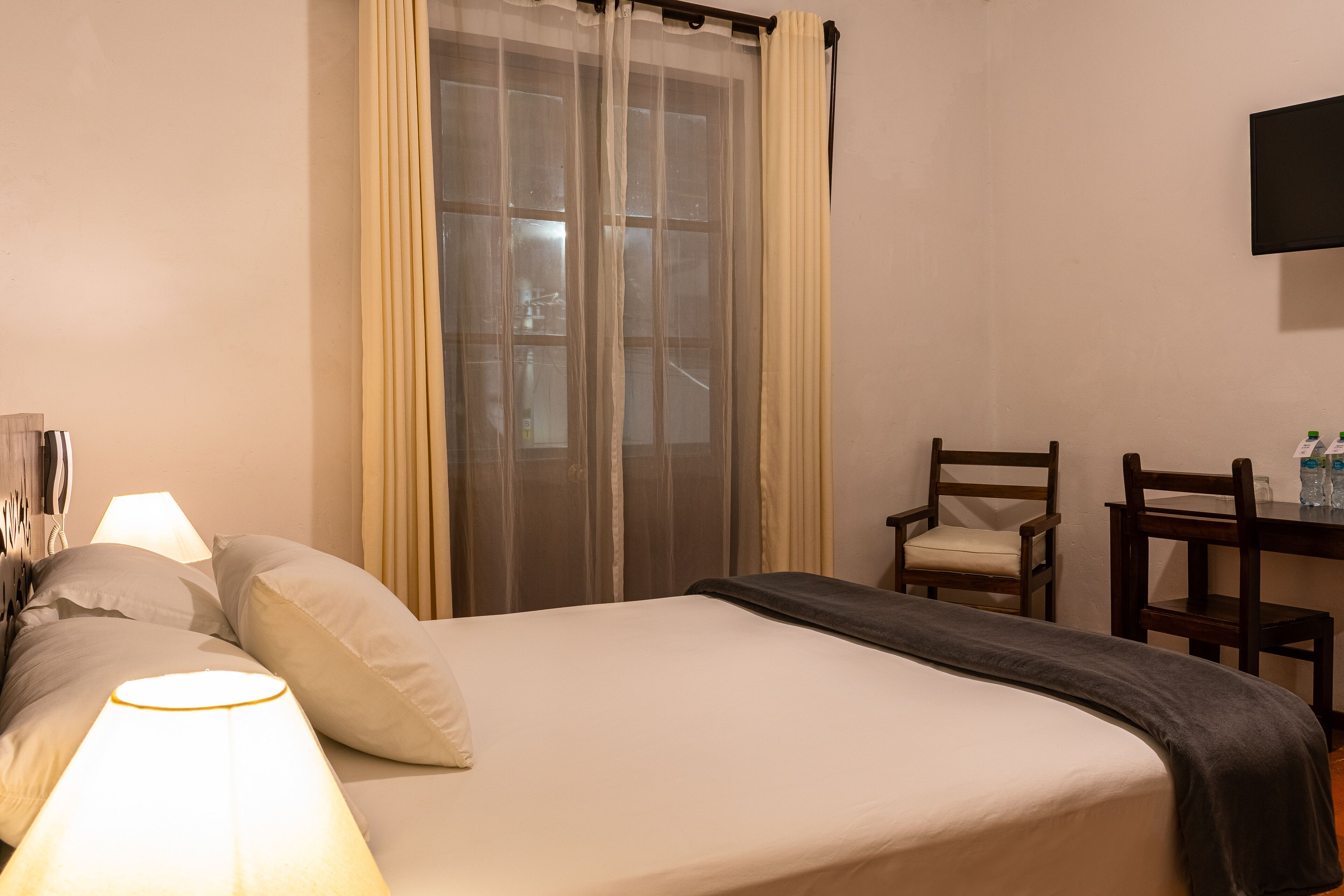 Quarto de hotel com cama, escrivaninha, cadeira, televisão e janela com cortinas.