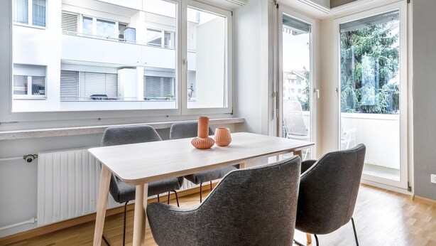 Un apartamento moderno con comedor que incluye una mesa blanca y sillas grises, ventanas amplias con persianas y vista a un edificio residencial.