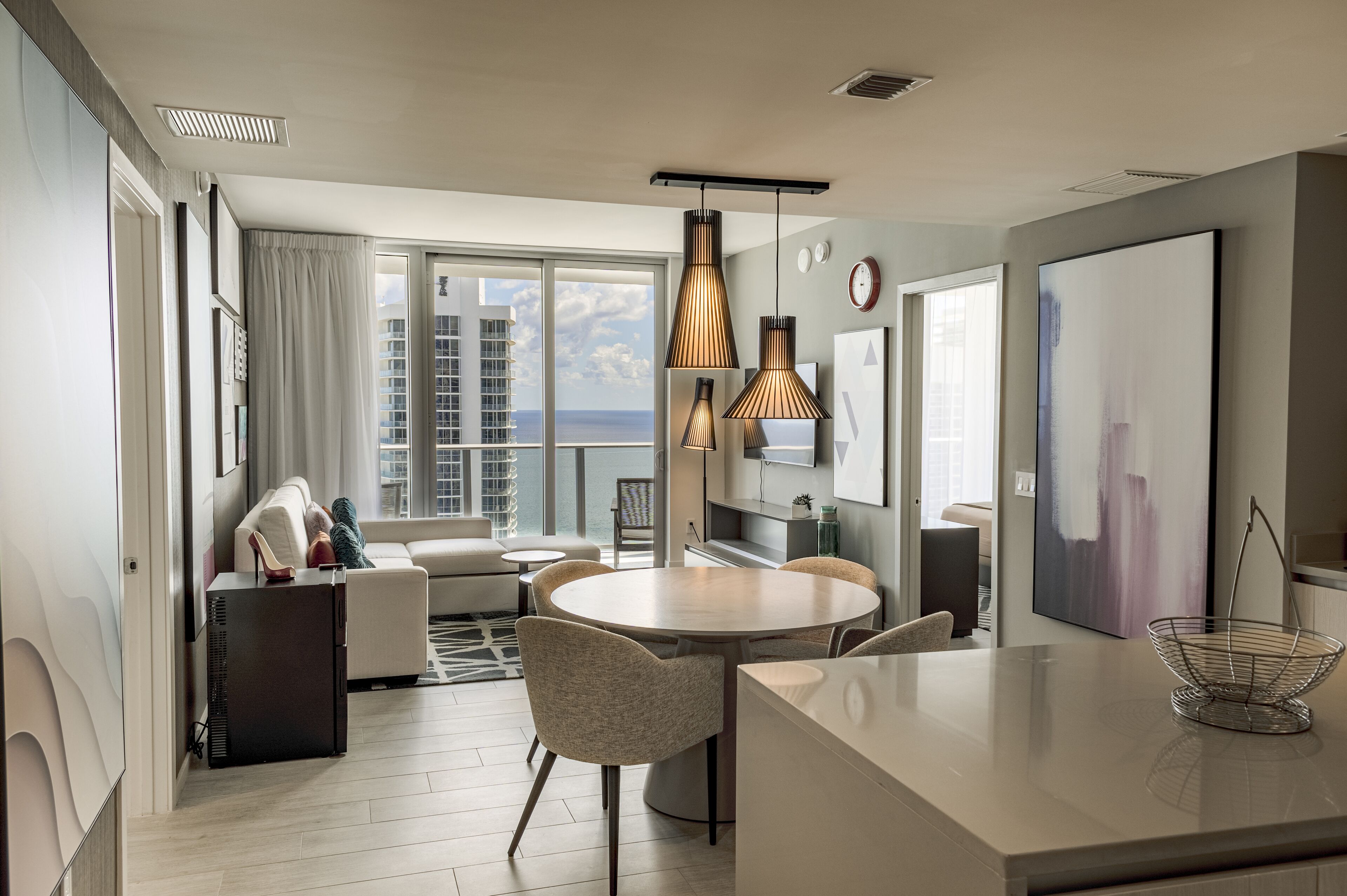 A modern hotel room with a dining area, a sofa, a TV, and a large window with a city view.