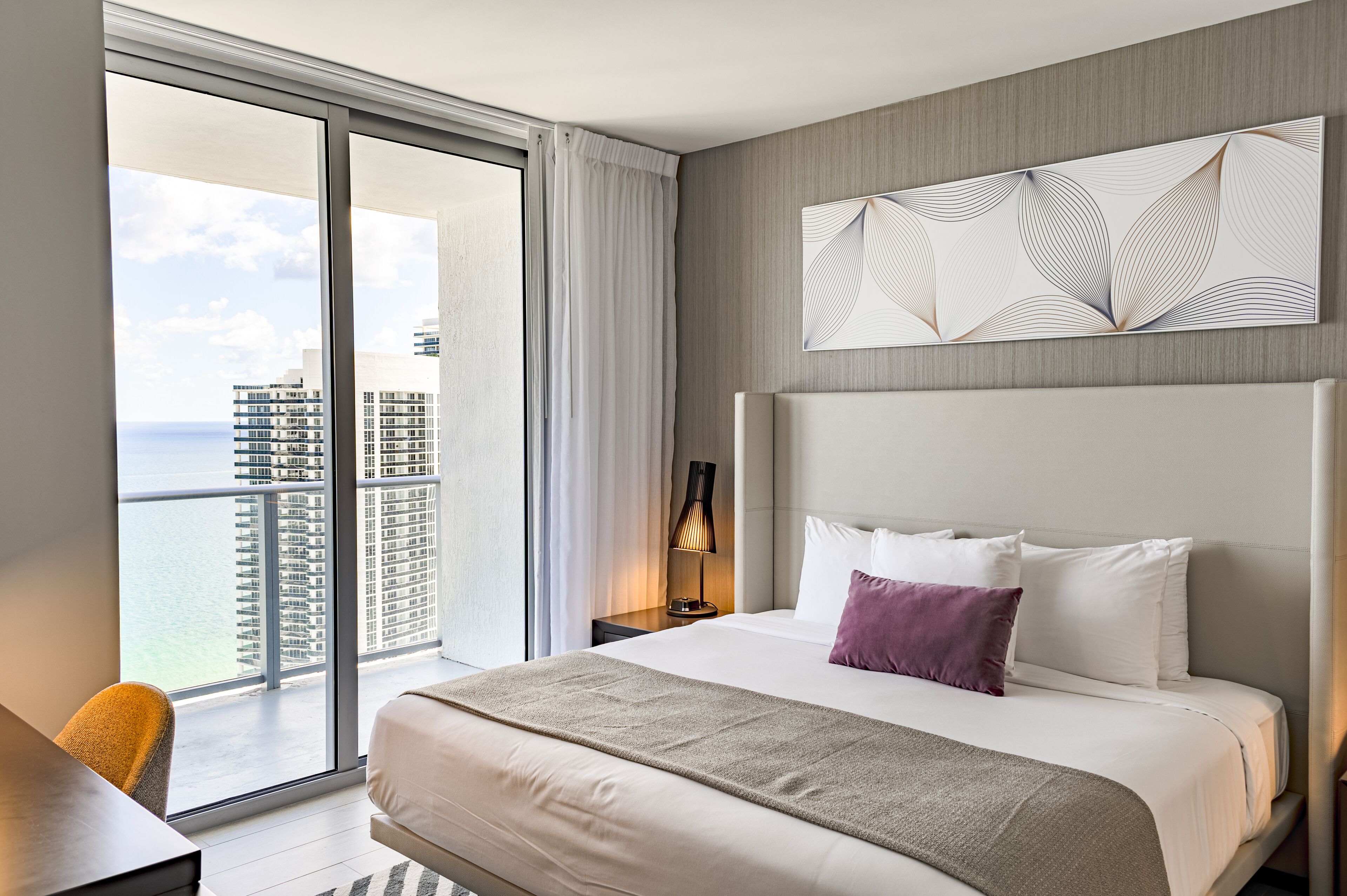 A hotel room with a large bed, a nightstand, a lamp, and a view of the ocean and buildings.