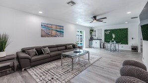 A modern living room with a sectional sofa, a glass coffee table, and a patterned rug. There is a dining area with a green wall and a bar counter in the background.