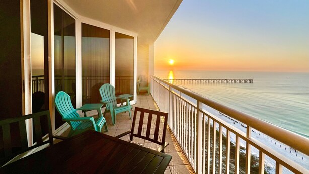 A balcony with a view of the sunset over the ocean, featuring a wooden table and two green chairs. - Updated 2BR/2BA, 14th Floor, Gulf & Sound Views, Seasonal Beach Service Included (Navarre Beach)