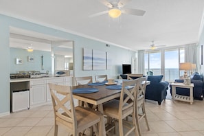 A spacious living area with a dining table, chairs, and a TV. There is a balcony with a view of the ocean.