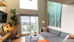 A modern living room with a grey sofa, a coffee table, a large window with a view, a potted plant, and a painting on the wall.