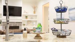 A kitchen counter with a stainless steel faucet, a soap dispenser, a dish soap bottle, and a three-tiered wire basket with snacks. - Upscale in Uptown West 4/2/2 (Lubbock)