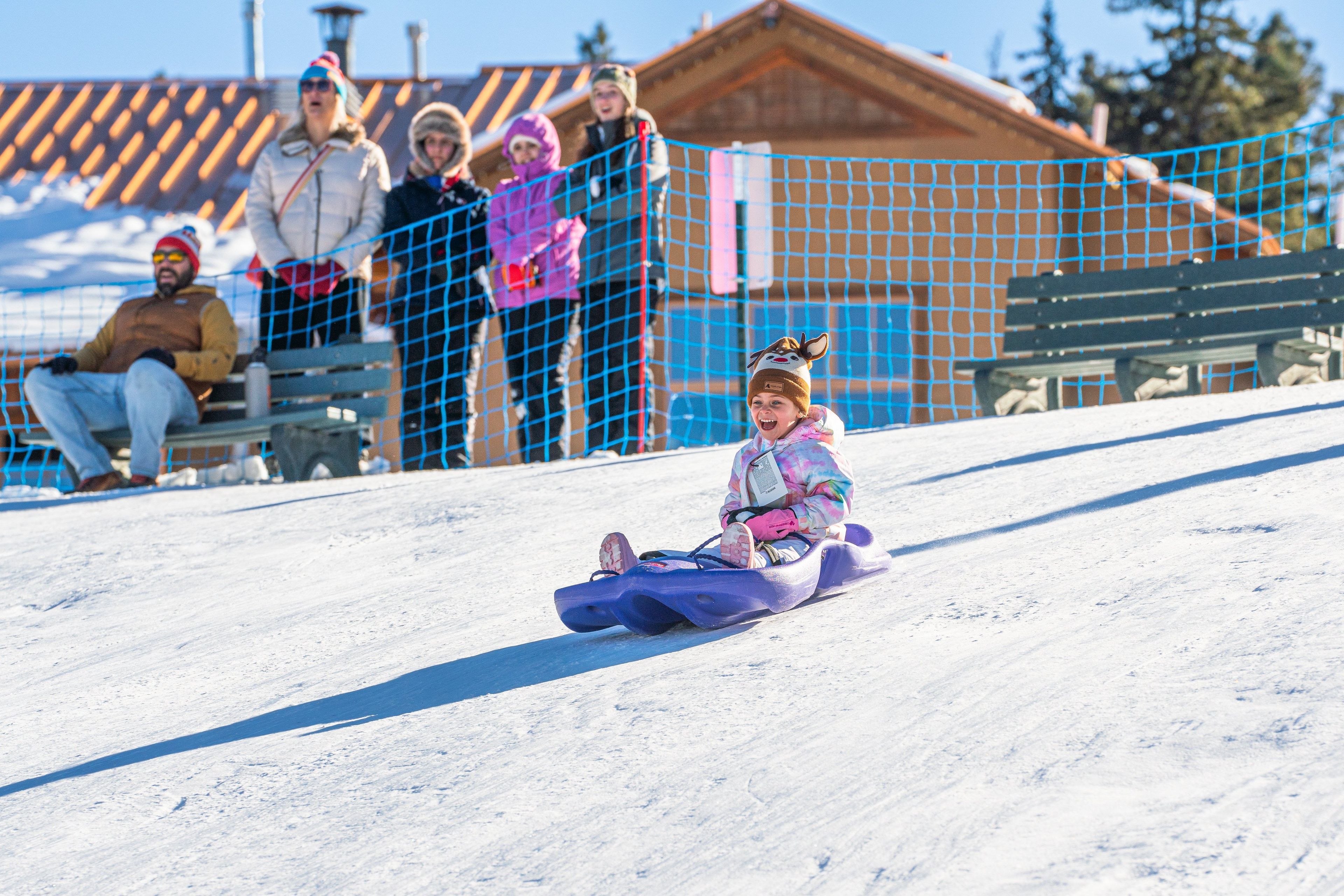 A snowy outdoor area with a sledding slope, spectators, and a building in the background.