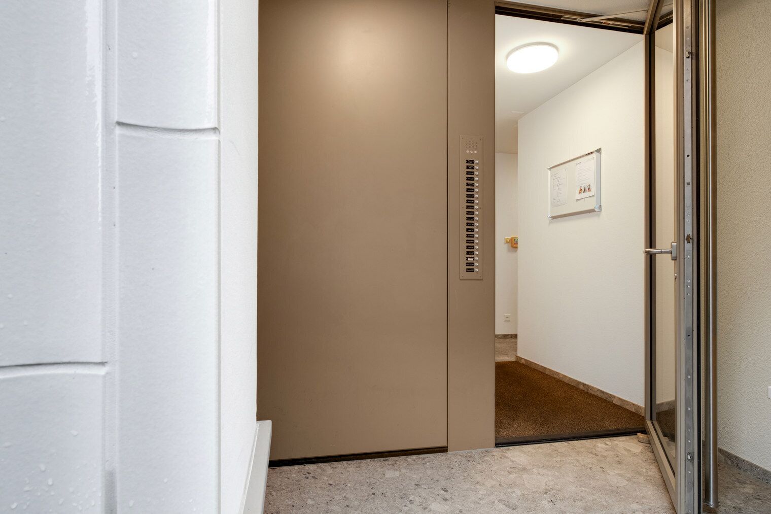 Un couloir étroit mène à une pièce par une porte. Le couloir a un plancher et des murs de couleur claire, et la porte est dotée d’une poignée métallique.