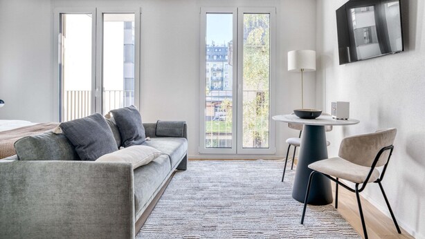 A modern hotel room with a grey sofa, a small round table with a lamp, and a TV mounted on the wall. - Sunlit Wiedikon 1 Room w/ Storage & W/D, 1 min to Market, by Blueground (Zurich)
