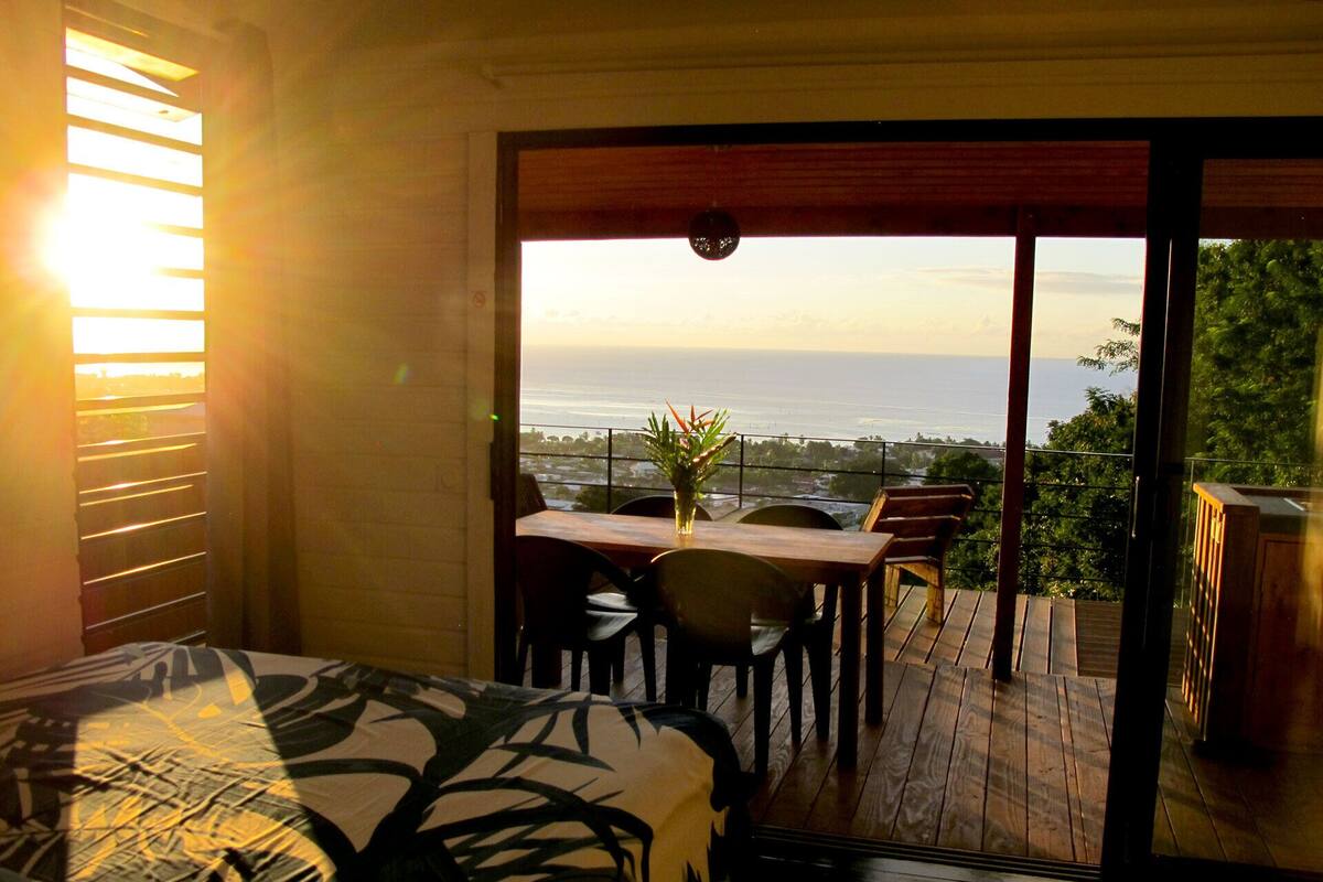 A bedroom with a bed, a wooden table, and chairs, and a balcony with a view of the ocean.