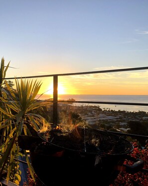 A balcony with a view of the sunset over the sea, a grill, and some plants.