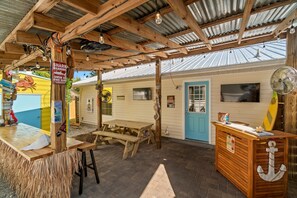 Un bar junto a la playa con una mesa de madera y bancos, una tabla de surf sobre el mostrador y un letrero de advertencia sobre tiburones.