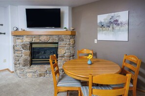 A dining area with a stone fireplace, a flat-screen TV mounted above it, and a painting on the wall. - Peacefield at The Hilltop Condo (Fish Creek)