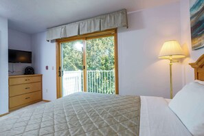 Ein Schlafzimmer mit Bett, Kommode, Fernseher, Lampe und einem Balkon mit Blick auf Bäume.