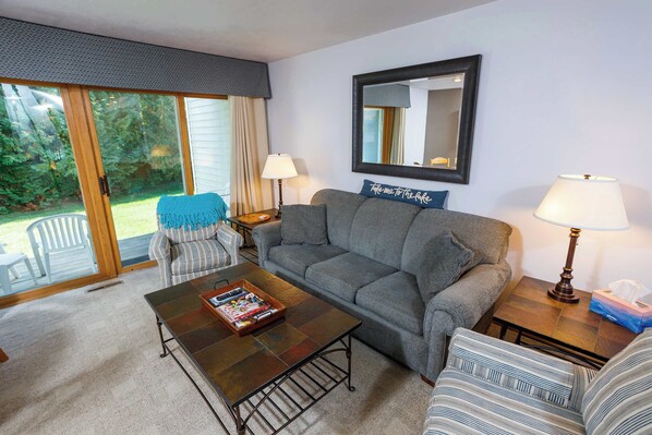 A living room with a gray sofa, striped armchair, and a coffee table with a magazine. There is a window with a view of greenery outside.