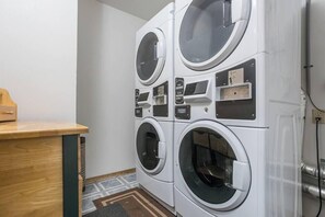 A laundry room with four stacked washing machines, a wooden counter, and a trash bin. - Peacefield at The Hilltop Condo (Fish Creek)