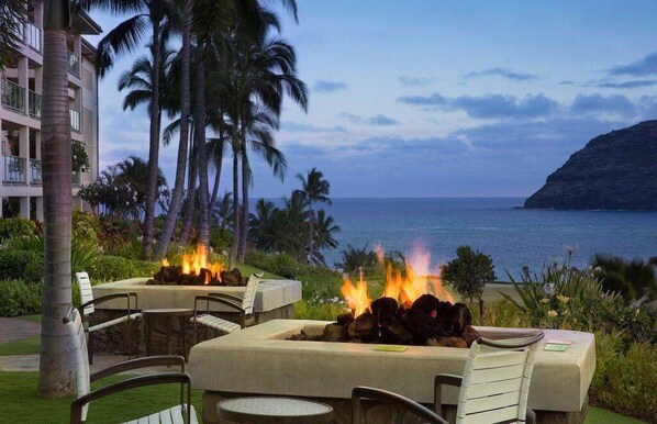 An outdoor seating area with a fire feature, overlooking a coastal view with palm trees and a building in the background.