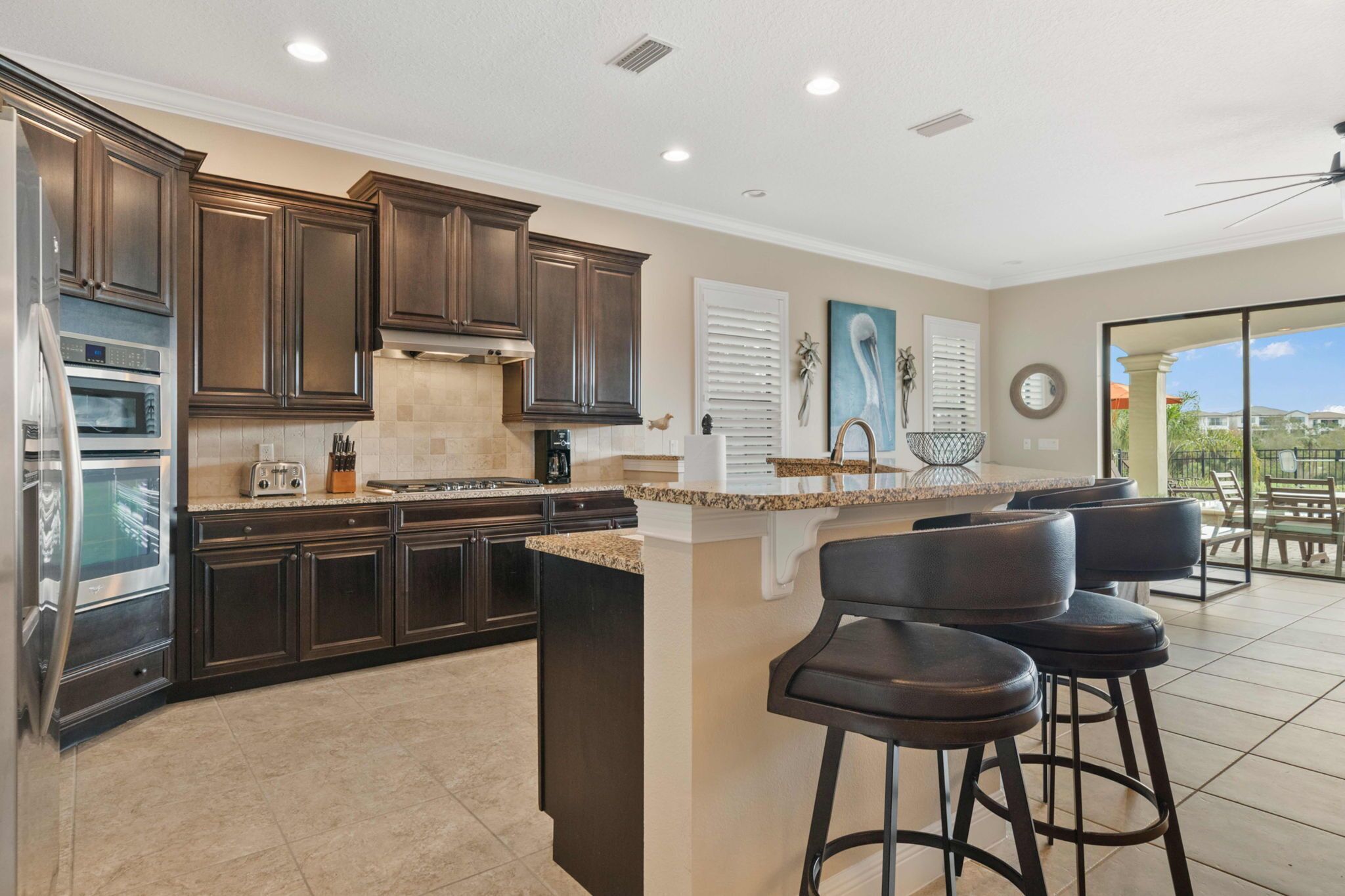 A modern kitchen with dark wood cabinets, a central island with bar stools, a built-in oven, and a large window overlooking a patio area.
