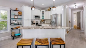 Una cocina moderna con armarios blancos, encimera de granito y electrodomésticos de acero inoxidable. Hay tres taburetes de madera en la isla.