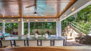 Un patio al aire libre con una mesa y sillas de madera, un ventilador de techo y vistas a un paisaje natural con una formación rocosa y escaleras.
