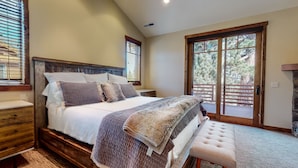 A bedroom with a large bed, wooden headboard, and a bench. There are windows with blinds, a fireplace, and a view of trees. - Graystone #2024 4 Bedroom Home by RedAwning (Mammoth Lakes)