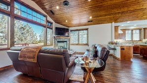 A spacious living room with a leather sofa, a glass coffee table, and a fireplace. Large windows offer views of trees. - Graystone #2024 4 Bedroom Home by RedAwning (Mammoth Lakes)