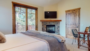 A bedroom with a large bed, a stone fireplace, a television above the fireplace, and a balcony with a wooden railing. - Graystone #2024 4 Bedroom Home by RedAwning (Mammoth Lakes)