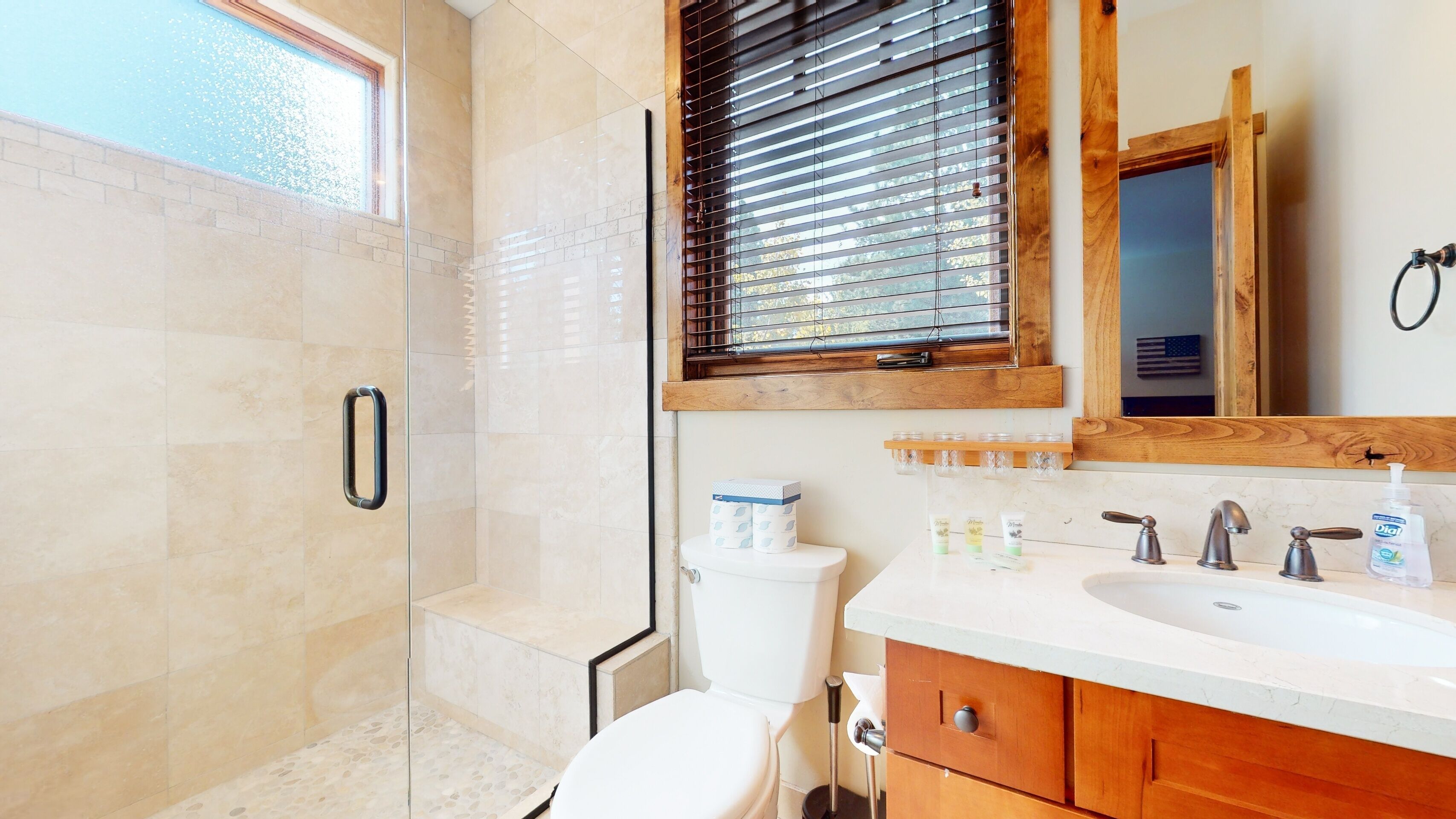 A bathroom with a glass shower enclosure, a toilet, a sink with a wooden vanity, and a window with blinds.