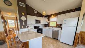 A kitchen with white cabinets, a central island, wooden chairs, a microwave, a refrigerator, and a window with a view. - The Dapper Duck - 4 BR Beach Cottage - Dogs Allowed (Southern Shores)