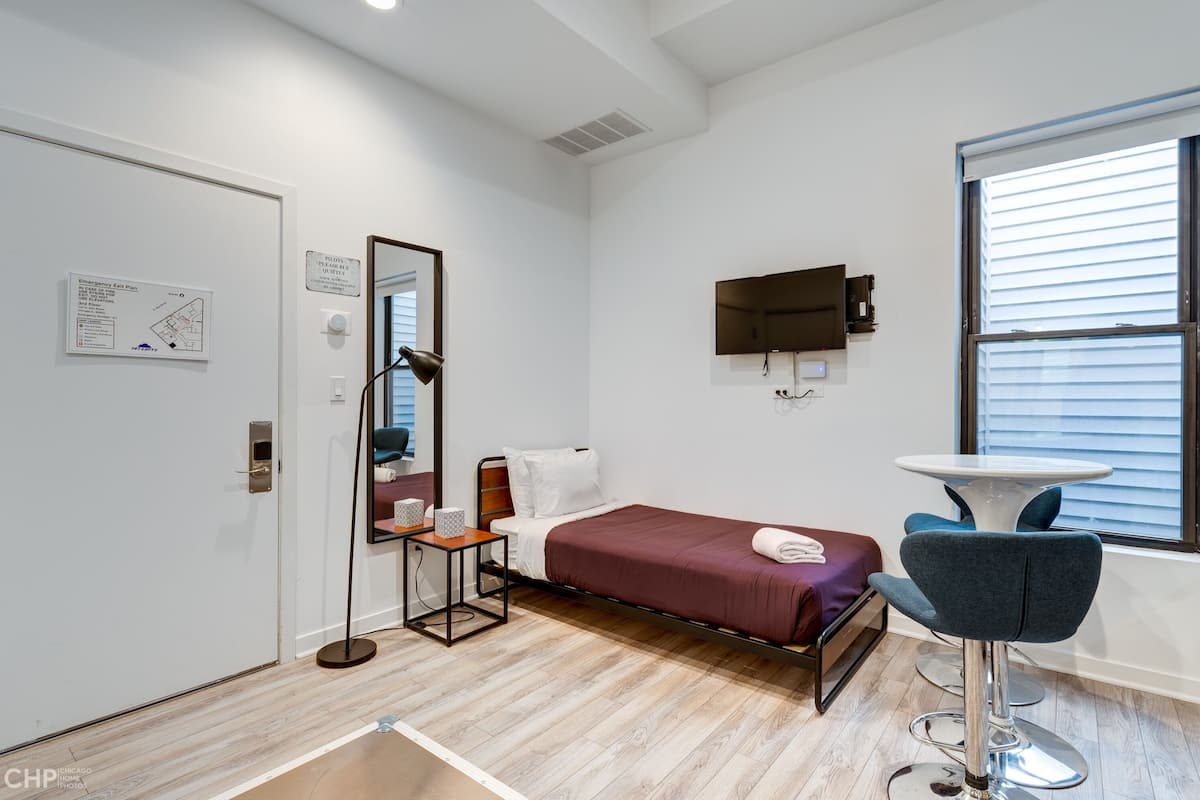 A modern hotel room with a single bed, a desk with a chair, a TV mounted on the wall, and a window with blinds.