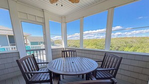 A covered outdoor patio with a round wooden table and chairs, large windows, and a view of a green landscape.