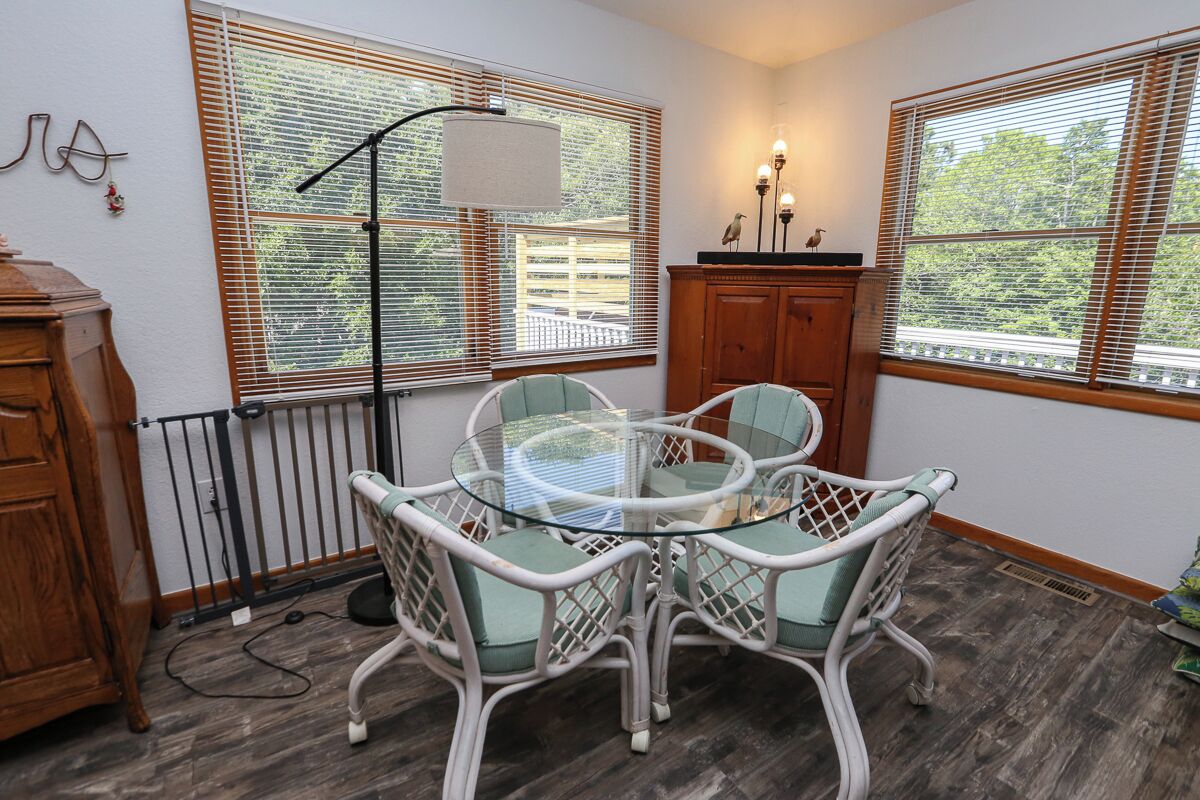 A dining area with a glass table and white wicker chairs, wooden cabinets, and a view through large windows.