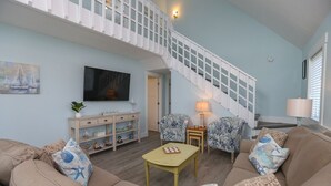 A spacious living room with a high ceiling, a staircase, and a television mounted on the wall. - Sandy Pointe -  4 BR Oceanside Home in Duck w/Private Pool, Hot Tub, Dogs Allowed (Duck)
