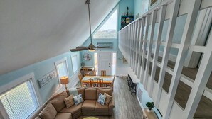 A spacious living area with a large L-shaped sectional sofa, a dining table, and a staircase leading to an upper level. - Sandy Pointe -  4 BR Oceanside Home in Duck w/Private Pool, Hot Tub, Dogs Allowed (Duck)