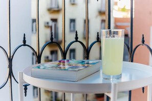 A table with a book and a glass of drink, set against a balcony railing with a building in the background.