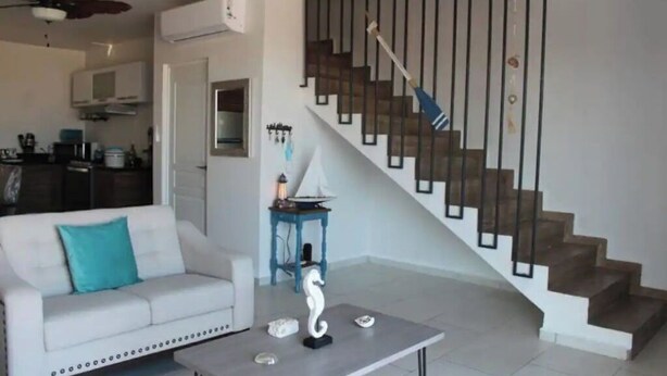 Un salon avec un canapé blanc, une table basse, un escalier doté d’une rampe en métal, et un coin cuisine à l’arrière-plan.