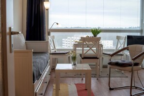 Une chambre moderne et bien éclairée, avec un lit, une petite table sur laquelle est posé un vase, deux chaises et une vue sur la ville grâce à de grandes fenêtres.