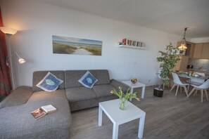 Un salon moderne avec un canapé gris, une petite table blanche et un coin repas agrémenté d’une plante et d’un panier de fruits.