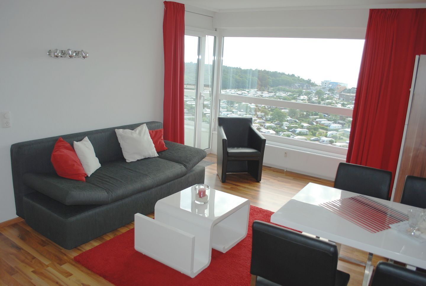 Een moderne woonkamer met een grijze bank, rode en witte kussens, een witte salontafel, en een eetgedeelte met een witte tafel en stoelen.