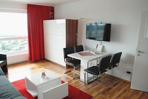 Een moderne woonkamer met een eethoek, een flatscreen televisie aan de wand en een groot raam met rode gordijnen.