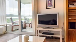 A living room with a flat-screen TV on a white stand, a glass door leading to a balcony with a chair, and a framed picture on the wall.