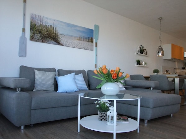 A modern living room with a grey sofa, a round coffee table with a vase of flowers, and a painting of a beach scene on the wall. - FB 05.10 - 4 person vacation apartment with modern furnishings, swimming pool and (Cuxhaven)