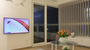 Un salon moderne avec un téléviseur à écran plat fixé au mur, une table basse en verre ornée d’un vase de fleurs et une porte-fenêtre coulissante donnant sur un balcon.