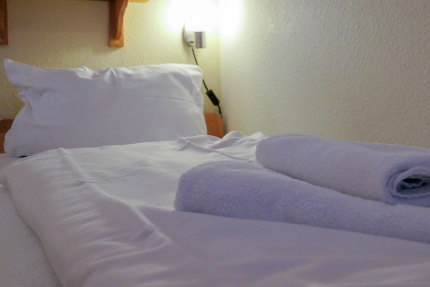 Una cama bien hecha con sábanas blancas y toallas enrolladas, una almohada blanca y un enchufe de pared.