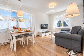 Ein modernes Wohnzimmer mit einer grauen Couch, einem Holzespeisetisch, weißen Stühlen, einem Fernseher und einer Stehlampe.