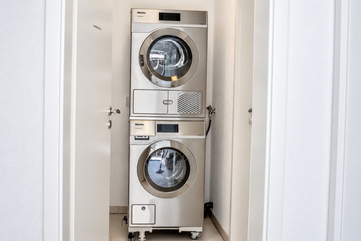 A compact, stainless steel, three-tiered, professional-grade, front-loading washing machine in a narrow hallway.