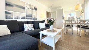 A modern living room with a sectional sofa, a white coffee table, and a dining area with white chairs. - DS2 - Nordic-style vacation rental near the beach for up to 4 people - House (Cuxhaven)