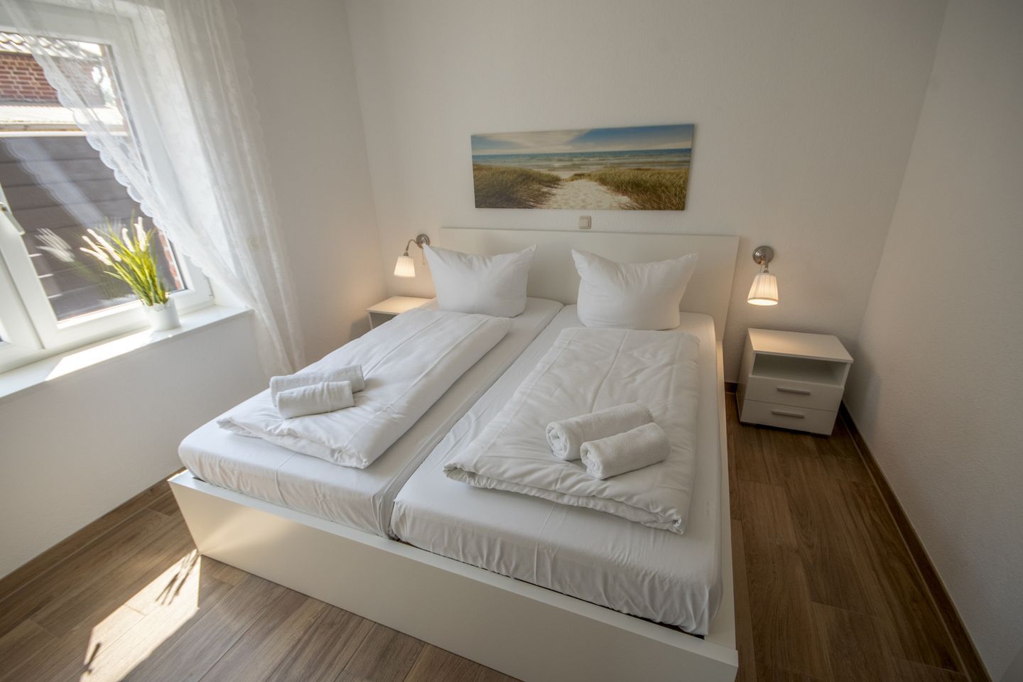Ein Doppelbett mit weißen Bettwäsche und Handtüchern, ein Nachttisch, ein Fenster mit Vorhängen und ein Bild an der Wand.