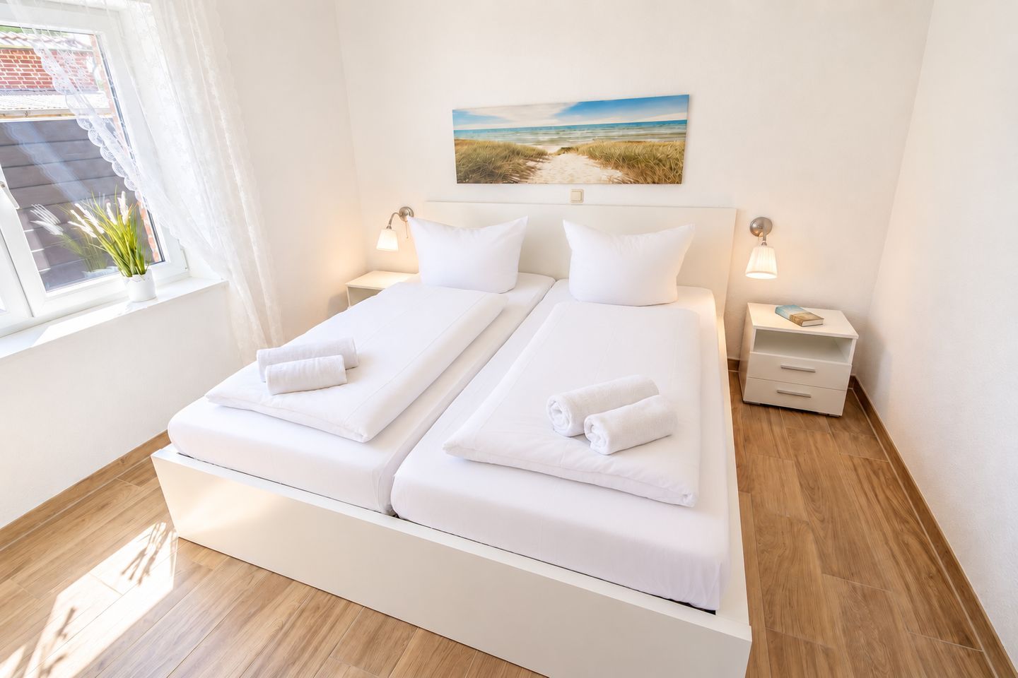 Un lit double avec du linge de lit blanc et deux oreillers, une table de chevet avec une lampe, et un tableau représentant une scène de plage accroché au mur.