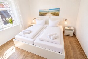 Una cama doble con sábanas blancas y dos almohadas, una mesita de noche con lámpara, y un cuadro de una playa en la pared.